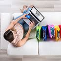 Above view of a woman sitting on a white couch holding a laptop with multi-colored shopping bags next to her.